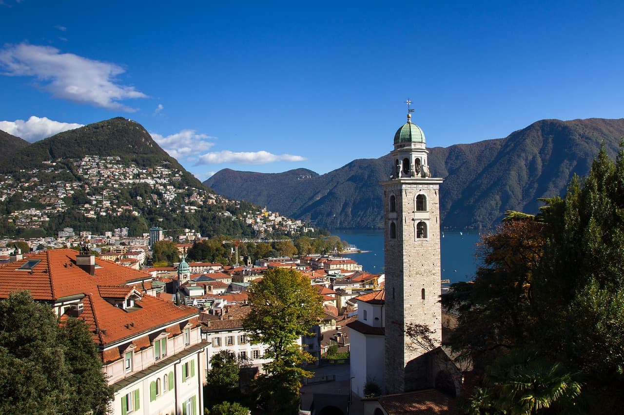Panorama der Altstadt von Lugano mit historischem Kirchturm, roten Dächern und Blick auf den Luganersee und die Berge im Tessin in der Schweiz.