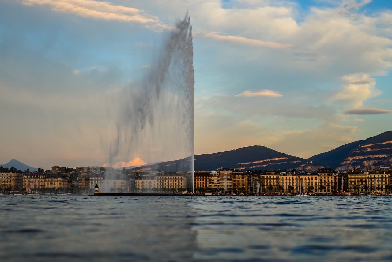 Berühmte Wasserfontäne Jet d’Eau in Genf am Genfersee mit Stadt und Bergen im Hintergrund bei Sonnenuntergang.